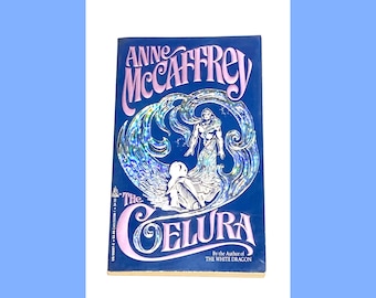 May include: A blue book cover with a holographic image of a person standing in water. The title of the book is "The Celura" by Anne McCaffrey. The text "By the Author of THE WHITE DRAGON" is printed below the title.