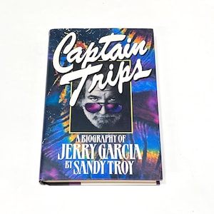 May include: A hardcover book titled "Captain Trips" with a portrait of Jerry Garcia. The cover features a tie-dye design in shades of blue, purple, and yellow. The book is a biography of Jerry Garcia by Sandy Troy.