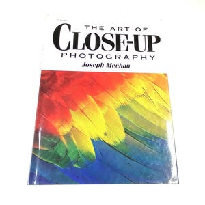 May include: A softcover book titled The Art of Close-Up Photography by Joseph Meehan. The white cover features black text and a vibrant, detailed photograph of colorful bird feathers in shades of red, yellow, green, and blue at the bottom.