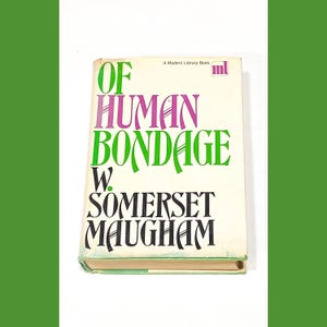 Puede incluir: Una edición de "Of Human Bondage" de W. Somerset Maugham, publicada por Modern Library. La cubierta del libro es blanca con texto verde y morado.