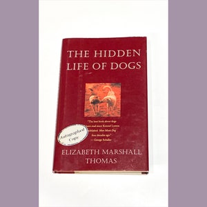 May include: A red hardcover book titled "The Hidden Life of Dogs" by Elizabeth Marshall Thomas. The book cover features a photo of two dogs interacting. The book is an autographed copy.
