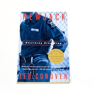 May include: A book cover with a blue background and a photo of a man in a blue uniform. The title of the book is "Newjack: Guarding Sing Sing" and the author is Ted Conover. The book is a winner of the National Book Critics Circle Award for Nonfiction.