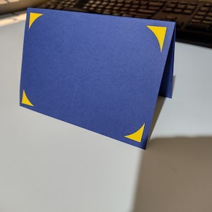 Op de afbeelding: Een blauwe kaart met gele driehoekige hoeken. De kaart is dubbelgevouwen en staat rechtop op een wit oppervlak.