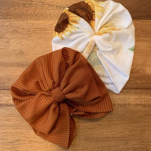 May include: Two fabric headbands, one with a sunflower print and the other a solid rust colour. Both headbands are tied in a knot at the top.