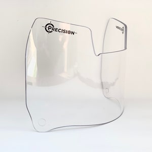 May include: A clear, curved plastic visor with the word "PRECISION-" printed in black. The visor has a rounded shape with a cut-out section and two small holes. The product is designed to protect the face.