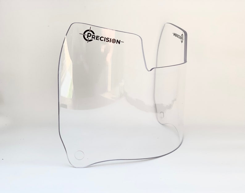 May include: A clear, curved plastic visor with the word "PRECISION-" printed in black. The visor has a rounded shape with a cut-out section and two small holes. The product is designed to protect the face.