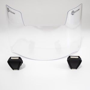 May include: A clear, curved visor with the word "PRECISION" printed on each side. Two black, geometric-shaped components are positioned below the visor. The items are set against a white background.