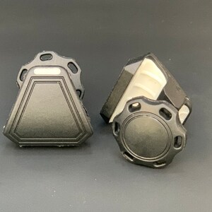 May include: Two black plastic objects with geometric shapes. One is a triangle with a decorative border and a rectangular top. The other is a circular shape with a ridged edge and a decorative border. Both have a matte finish.