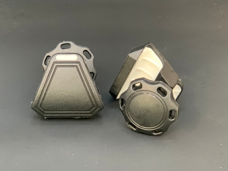 May include: Two black plastic objects with geometric shapes. One is a triangle with a decorative border and a rectangular top. The other is a circular shape with a ridged edge and a decorative border. Both have a matte finish.