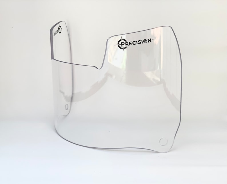 May include: A clear, curved visor with the word "PRECISION" printed in black. The visor has a rounded shape with a cut-out section and two small holes. The visor is made of transparent plastic.