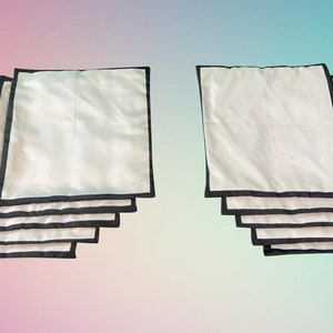 Op de afbeelding: Een set van tien witte kussenhoezen met zwarte bies. De hoezen zijn gestapeld in twee stapels van vijf.