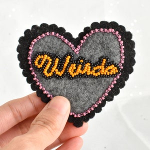 Puede incluir: Un parche de fieltro en forma de corazón con la palabra "Weirdo" bordada con cuentas naranjas y negras. El parche tiene un centro gris, un borde de cuentas rosas y un borde festoneado negro. El parche se sostiene en una mano.
