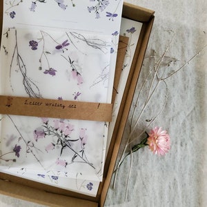 May include: A set of letter writing paper and envelopes with a floral design. The paper is white with a delicate pattern of purple and pink flowers. The envelopes are brown with a white floral design. The set is packaged in a brown cardboard box. The text "Letter writing set" is printed on the box.