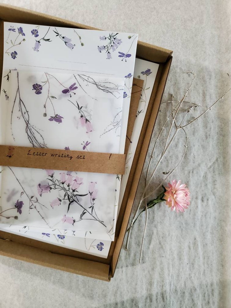 May include: A set of letter writing paper and envelopes with a floral design. The paper is white with a delicate pattern of purple and pink flowers. The envelopes are brown with a white floral design. The set is packaged in a brown cardboard box. The text "Letter writing set" is printed on the box.