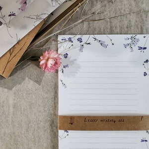 May include: A set of letter writing paper and envelopes with a floral design. The paper has a white background with blue flowers and the envelopes have a white background with purple flowers. The set is wrapped in brown paper with the text "Letter writing set" written on it.