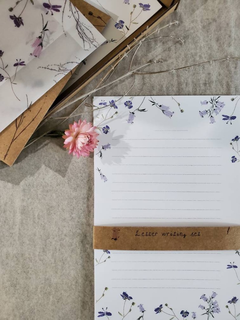 May include: A set of letter writing paper and envelopes with a floral design. The paper has a white background with blue flowers and the envelopes have a white background with purple flowers. The set is wrapped in brown paper with the text "Letter writing set" written on it.