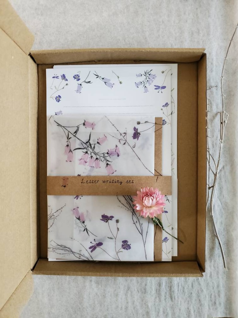 May include: A brown cardboard box with a white and purple floral patterned letter writing set inside. The set includes envelopes and writing paper. The box is open and a pink flower is sitting on top of the set.