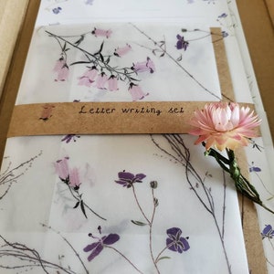 May include: A set of letter writing paper and envelopes with a floral design. The paper is white with a delicate pattern of pink and purple flowers. The envelopes are brown with a white floral design. The set is packaged in a brown cardboard box with the words "Letter writing set" printed on the front.