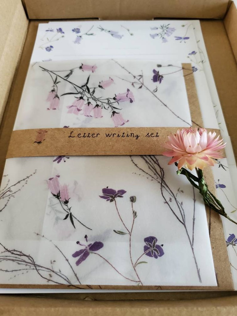 May include: A set of letter writing paper and envelopes with a floral design. The paper is white with a delicate pattern of pink and purple flowers. The envelopes are brown with a white floral design. The set is packaged in a brown cardboard box with the words "Letter writing set" printed on the front.