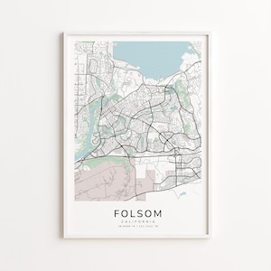 May include: Framed map print of Folsom, California, featuring a detailed street layout in black lines against a light background. The map includes a light blue water feature and green shaded areas. The city name and coordinates are printed at the bottom.