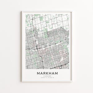 May include: A framed map print of Markham, Canada, featuring a detailed street grid and green shaded areas. The city name is displayed in a bold font at the bottom, with the coordinates below. The map is encased in a white frame.