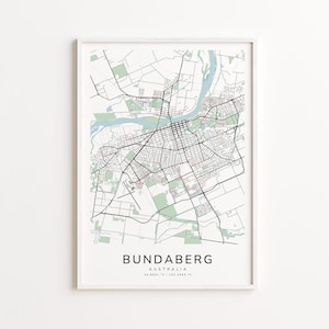 May include: A framed map print of Bundaberg, Australia, featuring a detailed street layout in black lines against a white background. Green areas represent parks and waterways. The city name and coordinates are printed at the bottom.