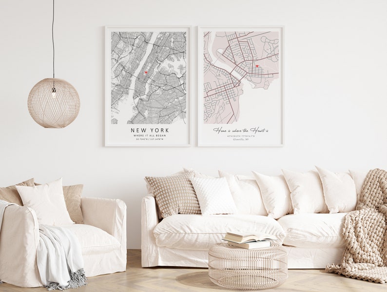 Op de afbeelding: Twee ingelijste kaarten, een van New York City en de andere van een kleinere stad. De kaart van New York City is zwart-wit met een rode pin die een locatie markeert. De kaart van de kleinere stad is roze met een rode pin die een locatie markeert. De tekst op de kaarten luidt "New York, Where it all began" en "Home is where the heart is".