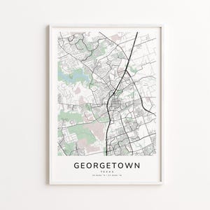 May include: A framed map print of Georgetown, Texas, in black and white, with green and pink accents. The city name is printed in a bold, sans-serif font at the bottom, with the coordinates below. The map is encased in a white frame.