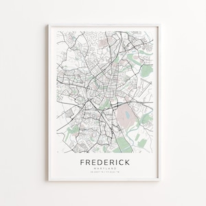May include: Framed map print of Frederick, Maryland, featuring a detailed black and white street layout with green and pink shaded areas. The city name is centered at the bottom, with coordinates below. The print is in a white frame.
