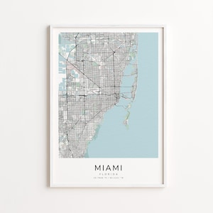 May include: Framed print of a Miami, Florida map. The map features a detailed street grid in gray and black, with a light blue area representing the ocean. The city name is printed in a bold font at the bottom.