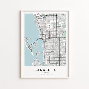 May include: A framed map print of Sarasota, Florida, featuring a detailed street grid and coastal areas in shades of black, white, and light blue. The city name is centered at the bottom, with coordinates below.