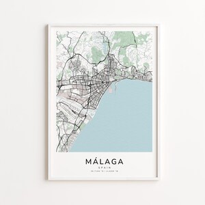 May include: A framed print of a map of Málaga, Spain. The map features a light blue body of water, green areas, and black roads and buildings. The city name "MÁLAGA" is printed in a bold, sans-serif font at the bottom, with the country name and coordinates below.