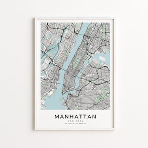 May include: A framed map print of Manhattan, New York, featuring a detailed street layout in black and gray against a light blue and pink background. The word "MANHATTAN" is printed in a bold, black font at the bottom, with the city's coordinates below.
