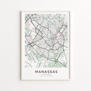 May include: Framed map print of Manassas, Virginia, showing a detailed street layout in black lines against a light background. The city name and coordinates are printed at the bottom. The frame is white.
