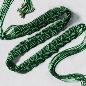 May include: A green braided cord belt with gold beads on the ends. The belt is made of multiple strands of cord that are woven together to create a decorative pattern.