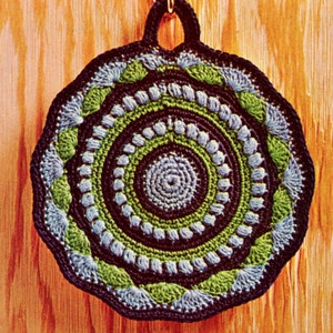 May include: A round, crocheted potholder with a dark blue border and hanging loop. The design features concentric circles in shades of blue, green, and dark blue. The potholder is hanging on a wooden surface.