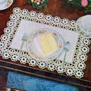 Puede incluir: Un mantel individual rectangular de encaje blanco con un delicado borde de ganchillo floral. Centrado sobre el mantel hay un plato de cerámica con motivos florales que contiene una porción de pastel de capas amarillo y rosa, con cubiertos.