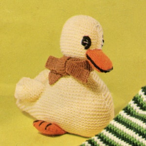May include: A crocheted duck toy with a brown scarf. The duck is white with orange feet and a black outlined orange beak. The duck is sitting on a green and white striped surface.