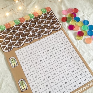 Può includere: Un tabellone di conteggio a forma di casetta di pan di zenzero con i numeri da 1 a 100 stampati su di esso. Il tabellone è marrone con glassa bianca e ha due finestre. Ci sono gettoni di conteggio colorati sparsi intorno al tabellone.