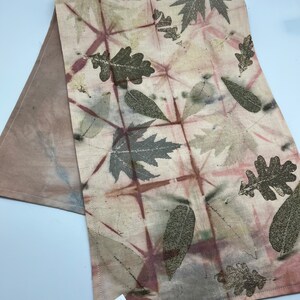 May include: A beige linen table runner with a leaf pattern in shades of brown and pink. The runner is decorated with a grid pattern and has a business card attached with the text "Wildwood Gallery, Lori Kovash, 520-334-5528, lori.kovash@yahoo.com".