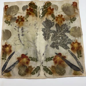 May include: A square, off-white textile with a botanical print. The design features pressed leaves and flowers in shades of brown, green, and orange. The edges are finished with a tan, stitched border. The overall theme is nature and art.