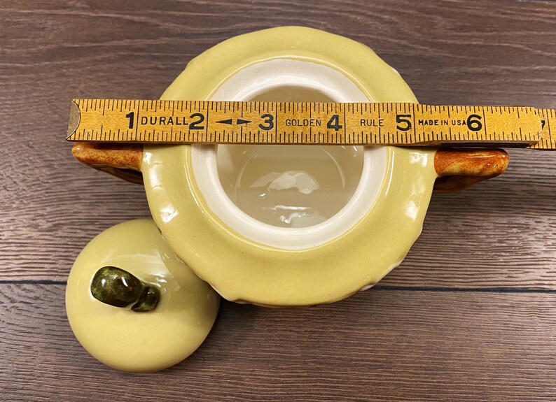 Op de afbeelding: Een gele keramische suikerpot met een deksel en een bruine handgreep. De pot heeft een wit interieur en een getextureerd exterieur. Het deksel heeft een bruine knop. De pot is 12,7 cm breed.
