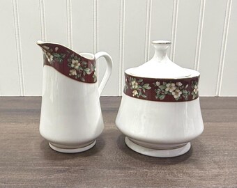 May include: A white ceramic creamer and sugar bowl set. The creamer has a handle and a curved rim. Both pieces feature a maroon band with a floral pattern of white and green flowers. The sugar bowl has a lid with a small knob.