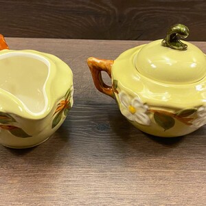 Op de afbeelding: Een gele keramische roomkan en suikerpot set met een bloemenmotief. De roomkan heeft een handvat en een tuit. De suikerpot heeft een deksel met een knop bovenop. Beide stukken hebben een wit bloemenmotief met groene bladeren en oranje accenten.