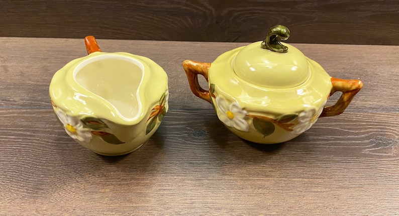 Op de afbeelding: Een gele keramische roomkan en suikerpot set met een bloemenmotief. De roomkan heeft een handvat en een tuit. De suikerpot heeft een deksel met een knop bovenop. Beide stukken hebben een wit bloemenmotief met groene bladeren en oranje accenten.