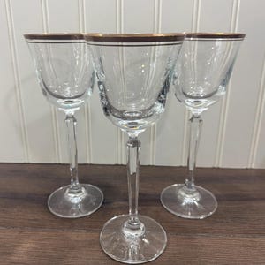 May include: Three clear glass cordial glasses with gold rims. The glasses have a slender stem and a round base. The glasses are arranged on a dark wood surface.