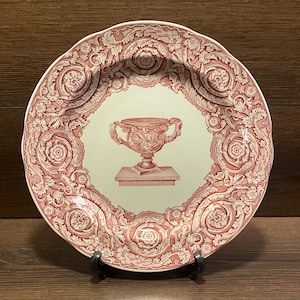 May include: A red and white ceramic plate with a decorative floral design. The center of the plate features a white ceramic urn with a floral design.