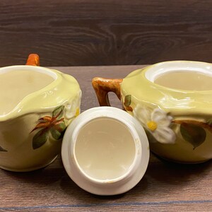 Op de afbeelding: Een vintage set van een keramische roomkan en suikerpot met een bloemenmotief. De roomkan en suikerpot zijn beide geel met witte bloemen en groene bladeren. De roomkan heeft een handvat en een tuit. De suikerpot heeft een deksel.