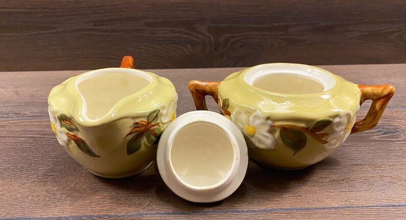 Op de afbeelding: Een vintage set van een keramische roomkan en suikerpot met een bloemenmotief. De roomkan en suikerpot zijn beide geel met witte bloemen en groene bladeren. De roomkan heeft een handvat en een tuit. De suikerpot heeft een deksel.