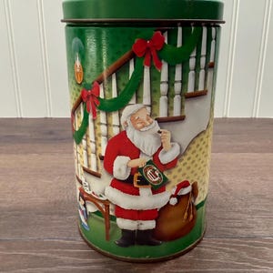 May include: A cylindrical Christmas tin with a green lid and base. The tin features a festive illustration of Santa Claus in his red suit, holding a treat, with a sack of gifts and a decorated staircase.